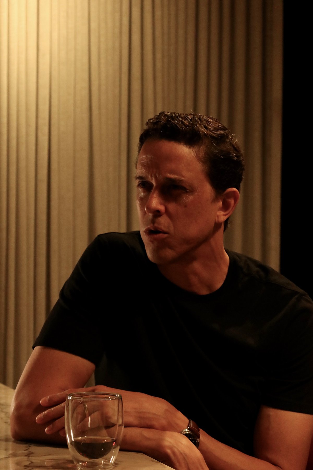 Man in black t-shirt sitting at table with glass of water, looking to the side against beige curtain background