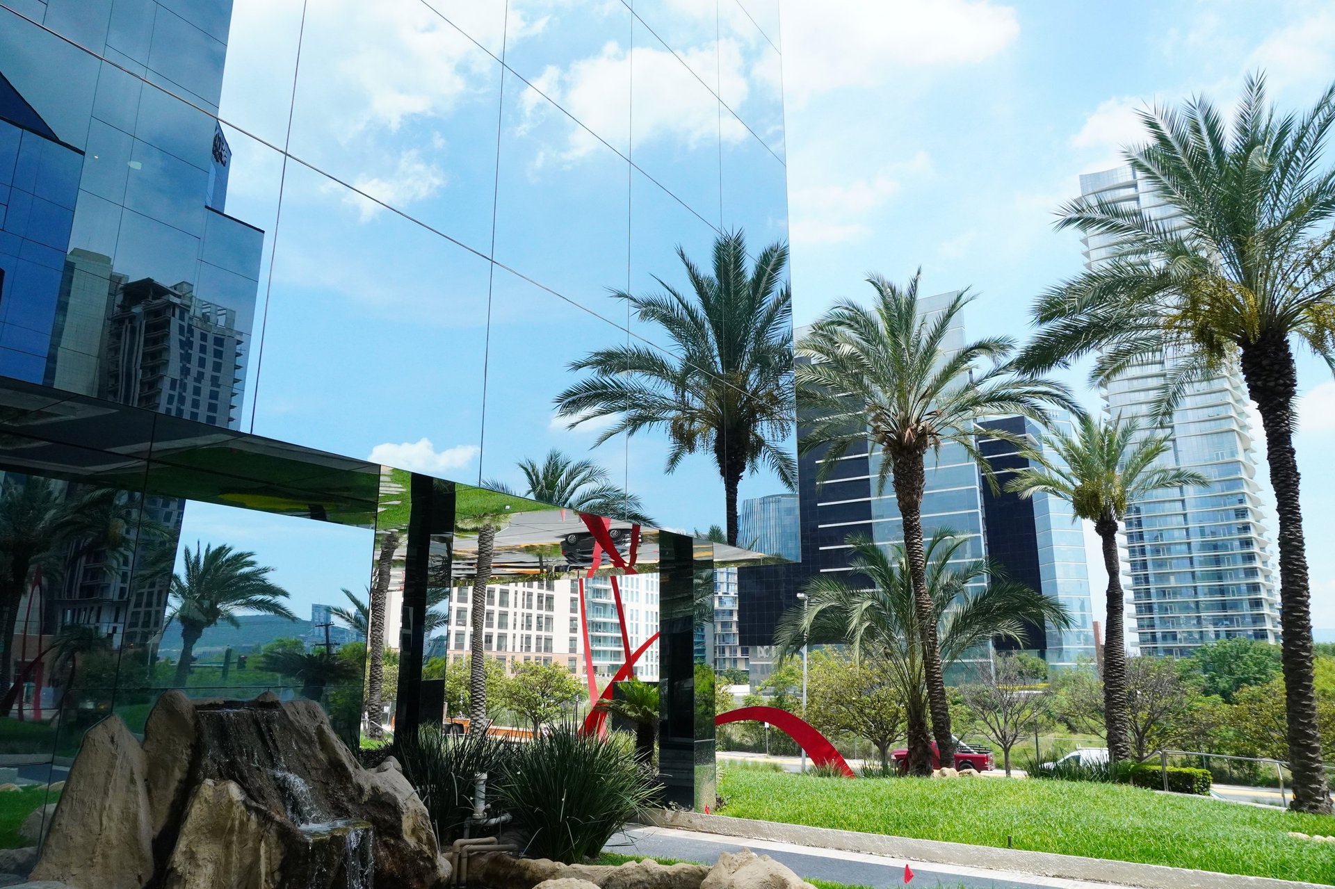 Urban park with palm trees, red slide playground, green lawn, and modern glass skyscrapers in background under blue sky