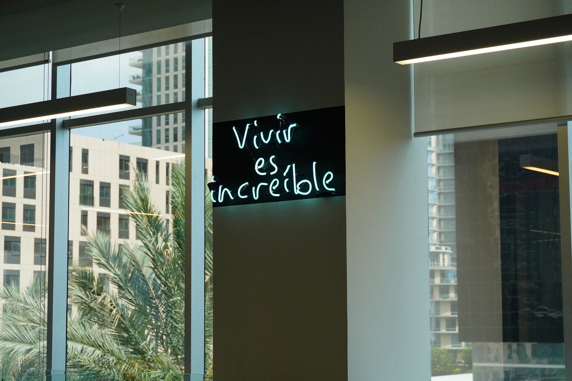 Modern office interior with green accent wall displaying white handwritten text reading Vivir es visible, large windows overlooking city buildings, and potted plant