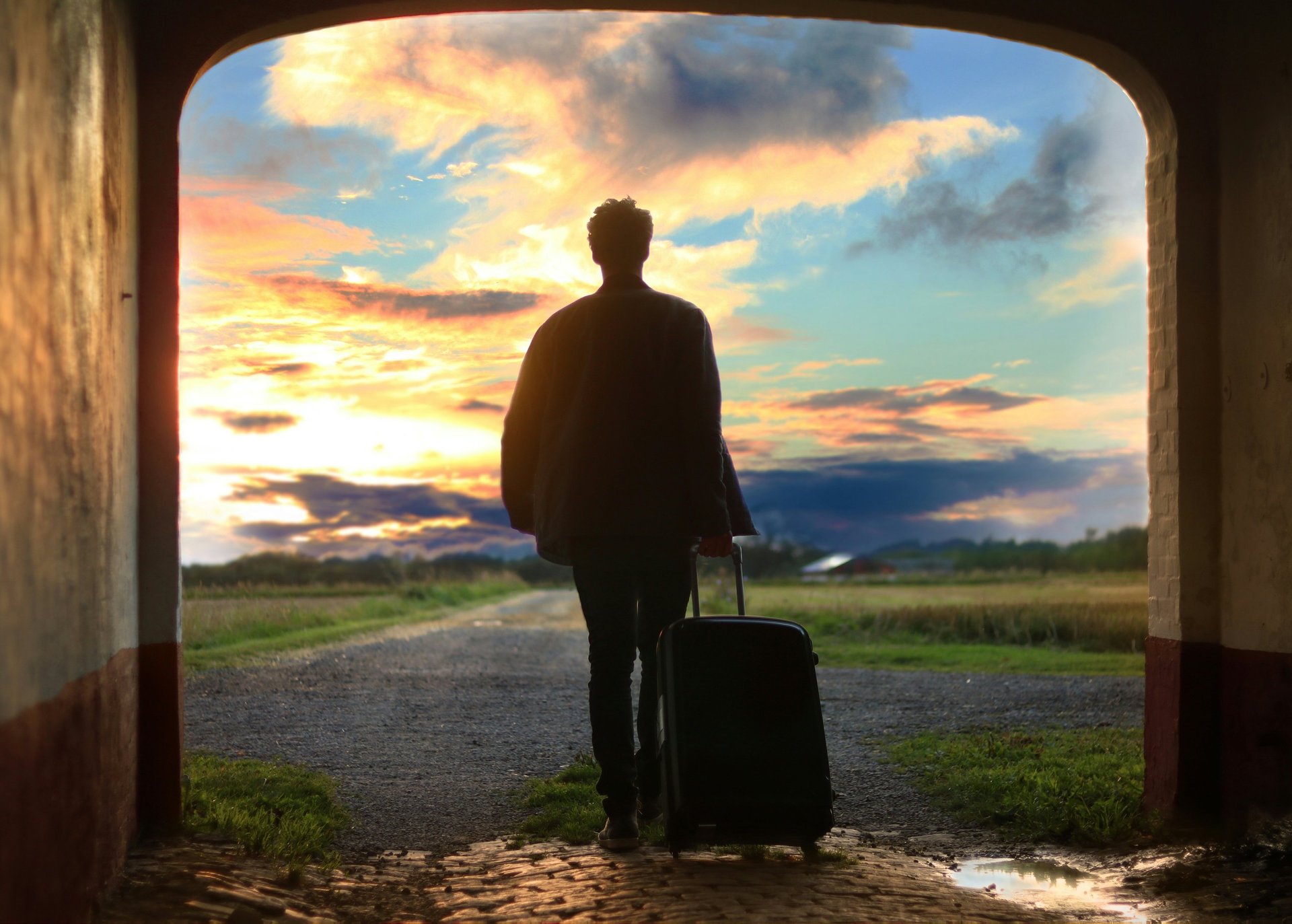 Silhouetted figure with suitcase standing in a doorway looking out at a scenic sunset landscape with green fields and mountains